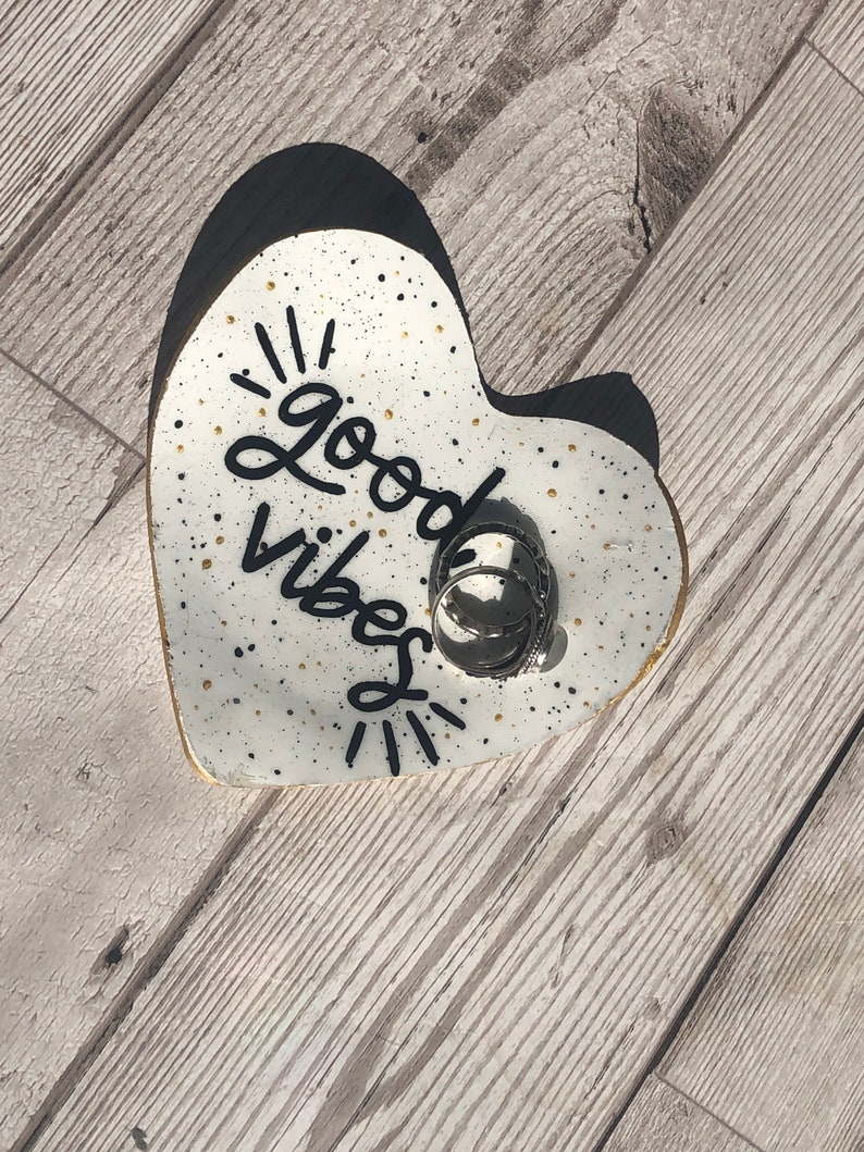 May include: White heart-shaped ceramic dish with black speckles and gold glitter. The dish has the words "Good Vibes" written in black cursive. There are several silver rings in the dish.