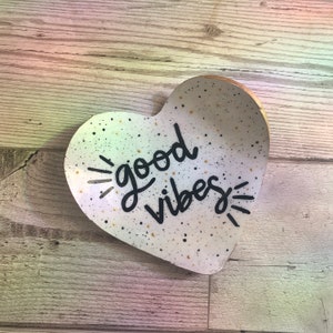 May include: White heart-shaped ceramic dish with black speckles and gold flecks. The dish has the words "good vibes" written in black cursive lettering.