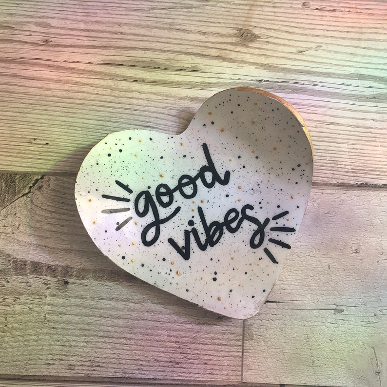 May include: White heart-shaped ceramic dish with black speckles and gold flecks. The dish has the words "good vibes" written in black cursive lettering.