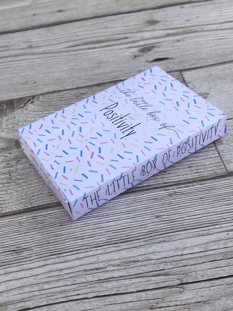 Puede incluir: Una caja blanca con un patr&oacute;n de confeti colorido. La caja tiene el texto "The Little Box of Positivity" en el lateral y "The little box of Positivity" en la parte superior.
