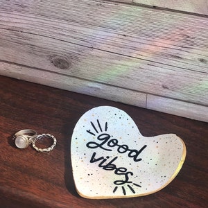 May include: White ceramic heart-shaped dish with gold speckles and black lettering that reads "good vibes".