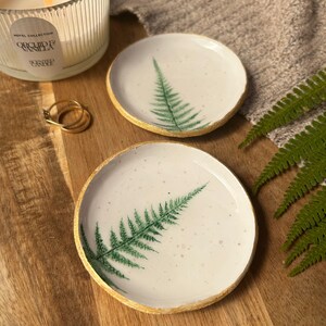 May include: Two round white ceramic trinket dishes featuring a green fern leaf print on the surface and a textured gold-painted rim. The dishes have a speckled finish and are displayed alongside a lit candle and two gold rings.