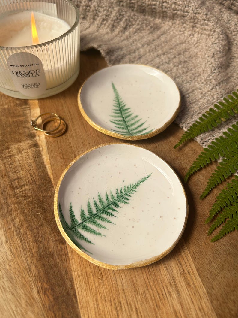 May include: Two round white ceramic trinket dishes featuring a green fern leaf print on the surface and a textured gold-painted rim. The dishes have a speckled finish and are displayed alongside a lit candle and two gold rings.