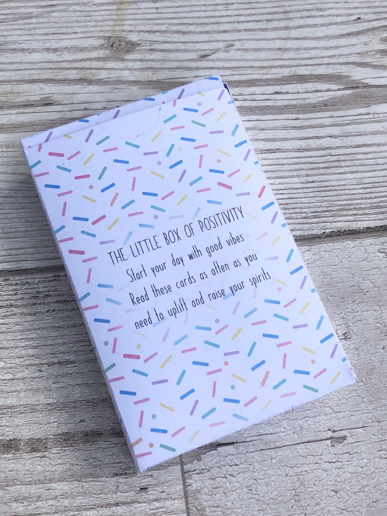 Puede incluir: Una peque&ntilde;a caja blanca con un patr&oacute;n de confeti colorido y el texto "THE LITTLE BOX OF POSITIVITY" impreso en la parte delantera. El texto debajo dice "Comienza tu d&iacute;a con buenas vibraciones. Lee estas tarjetas tan a menudo como necesites para animarte y levantar tu esp&iacute;ritu."