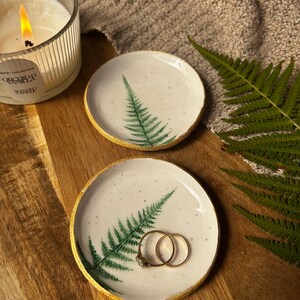 May include: Two round ceramic trinket dishes with a white speckled glaze, featuring a green fern leaf print and a metallic gold painted rim. One dish holds two gold rings, while the other is empty, showcasing the detailed botanical design.