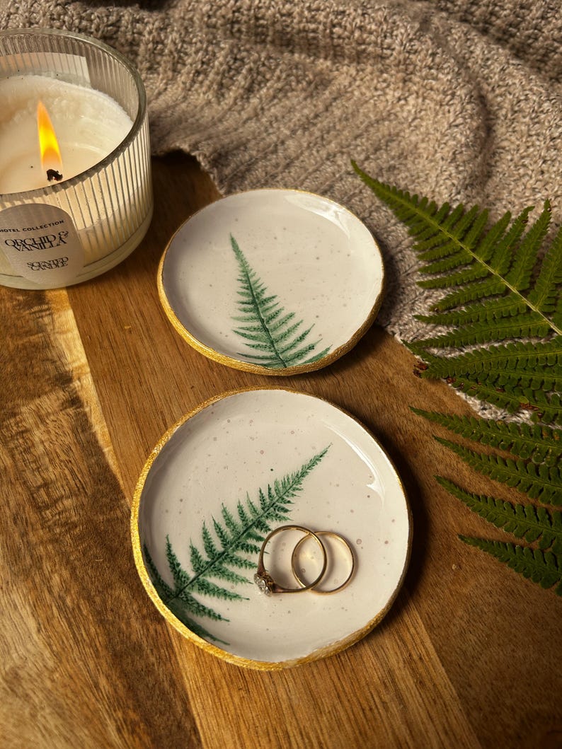 May include: Two round ceramic trinket dishes with a white speckled glaze, featuring a green fern leaf print and a metallic gold painted rim. One dish holds two gold rings, while the other is empty, showcasing the detailed botanical design.