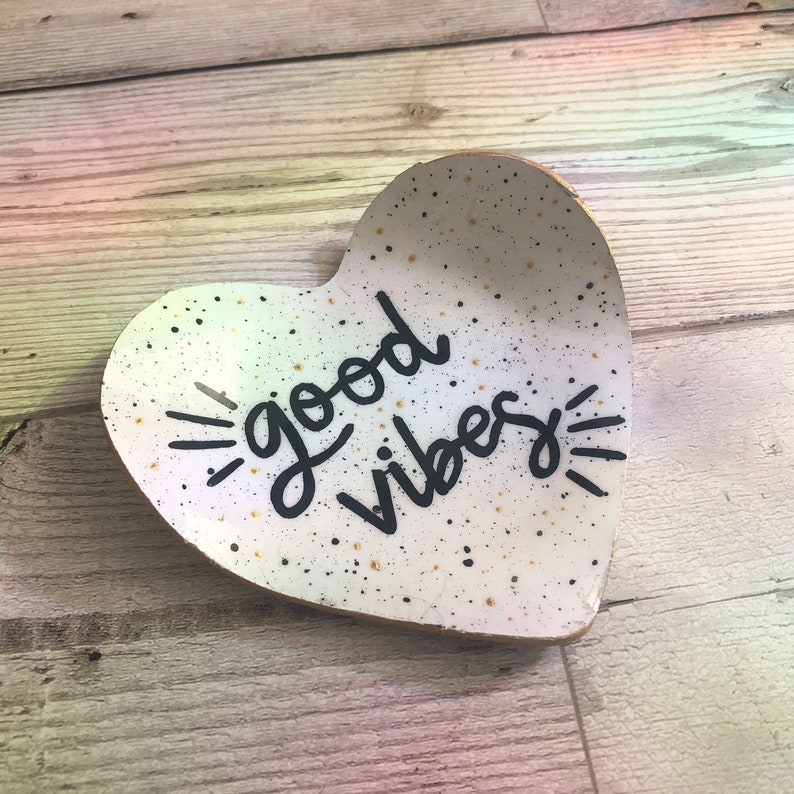 May include: White heart-shaped ceramic dish with black speckles and gold glitter. The dish has the words "good vibes" written in black cursive.