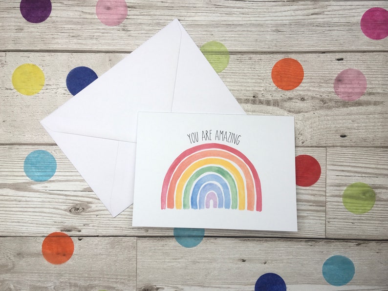 May include: A white card with a rainbow watercolour illustration and the text "You are amazing". The card is sitting on a white wooden surface with colourful polka dots.