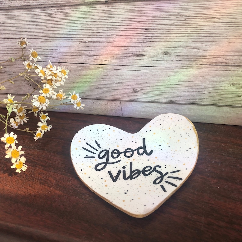 May include: White heart-shaped ceramic dish with black speckles and gold flecks. The dish has the words "good vibes" written in black cursive lettering.