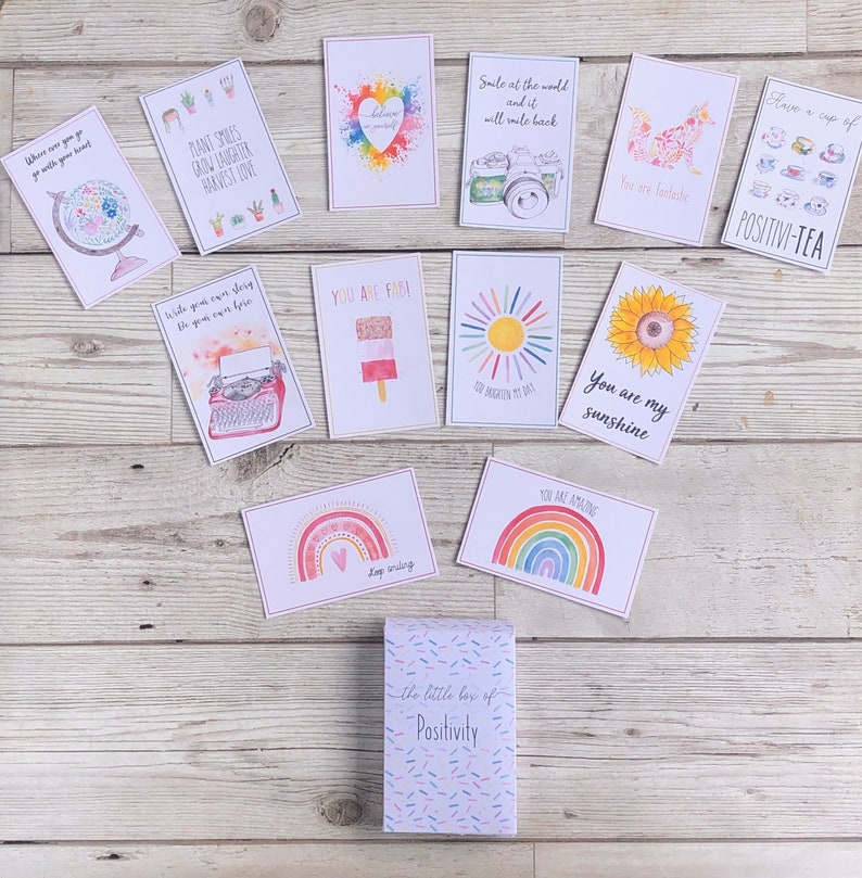Puede incluir: Una peque&ntilde;a caja con fondo blanco y estampado de confeti de colores. La caja est&aacute; etiquetada como "the little box of Positivity". La caja est&aacute; rodeada de 12 tarjetas con mensajes positivos e ilustraciones coloridas. Las tarjetas presentan arco&iacute;ris, un sol, una m&aacute;quina de escribir, una taza de t&eacute;, un globo terr&aacute;queo y un girasol.