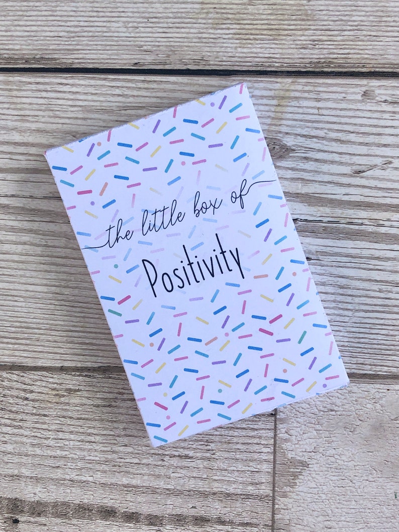 Puede incluir: Una peque&ntilde;a caja blanca con un patr&oacute;n de confeti colorido y el texto "the little box of Positivity" impreso en la parte delantera.