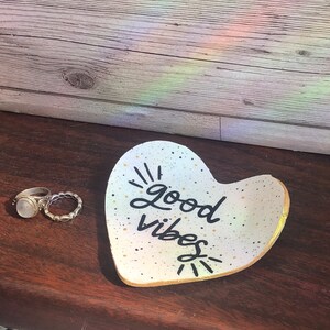 May include: White heart-shaped ceramic dish with gold speckles and black lettering that reads "good vibes".