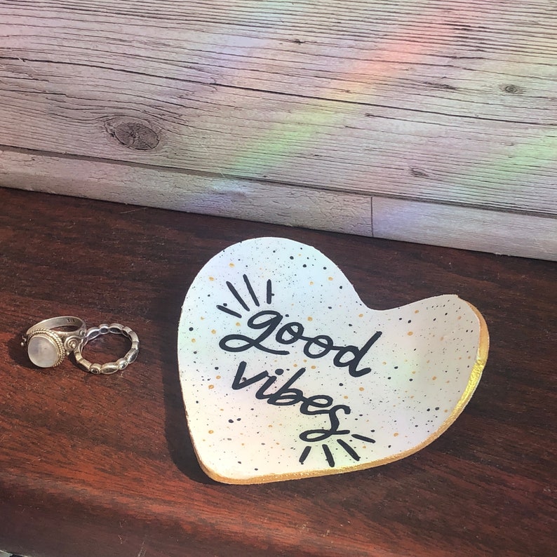 May include: White heart-shaped ceramic dish with gold speckles and black lettering that reads "good vibes".