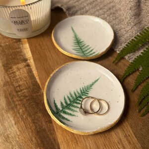 May include: Two round white ceramic trinket dishes featuring a green fern leaf print and gold-painted edges. One dish holds two gold rings. The dishes have a speckled finish and are placed on a wooden surface near a lit candle and a fern frond.