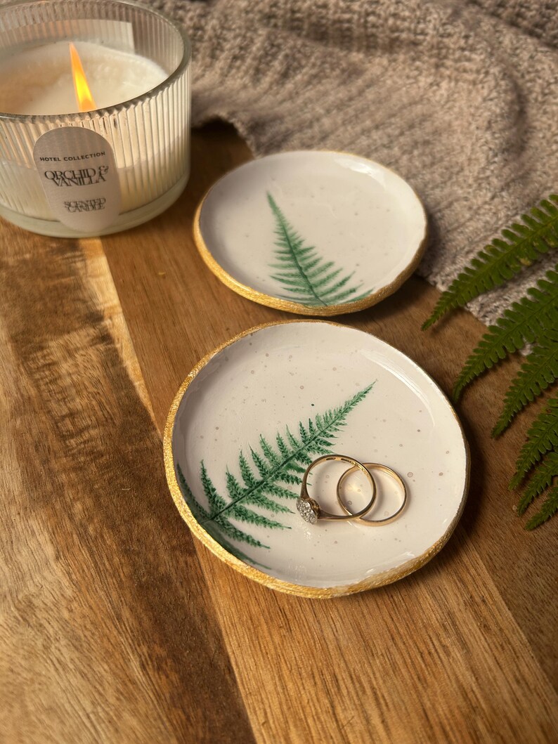 May include: Two round white ceramic trinket dishes featuring a green fern leaf print and gold-painted edges. One dish holds two gold rings. The dishes have a speckled finish and are placed on a wooden surface near a lit candle and a fern frond.