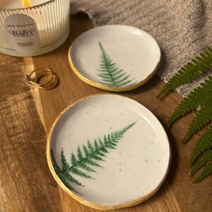 May include: Two round white ceramic trinket dishes featuring a green fern leaf print and gold-painted edges. The dishes have a speckled surface texture and are displayed alongside a ribbed glass candle with a lit wick and two gold rings.