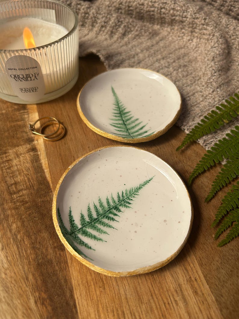 May include: Two round white ceramic trinket dishes featuring a green fern leaf print and gold-painted edges. The dishes have a speckled surface texture and are displayed alongside a ribbed glass candle with a lit wick and two gold rings.