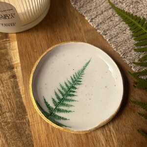 May include: A round, white ceramic trinket dish featuring a hand-painted green fern leaf design on its surface. The dish has a speckled texture and a metallic gold-painted rim, providing a distinct contrast against the white base of the object.