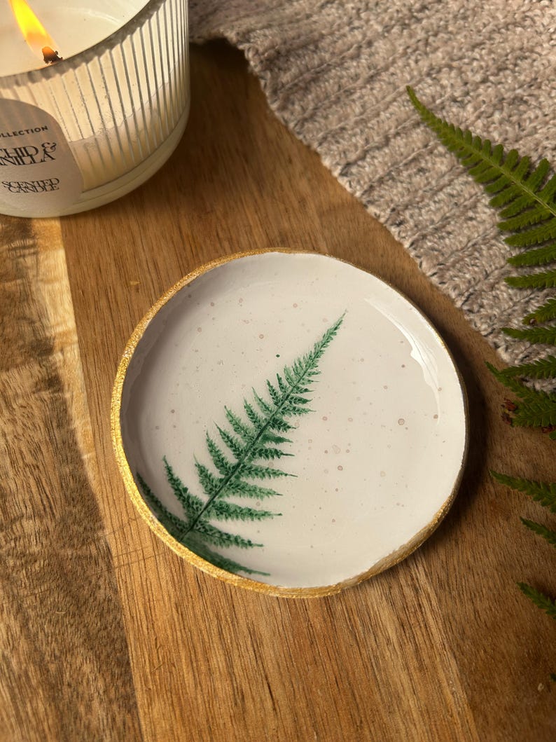 May include: A round, white ceramic trinket dish featuring a hand-painted green fern leaf design on its surface. The dish has a speckled texture and a metallic gold-painted rim, providing a distinct contrast against the white base of the object.