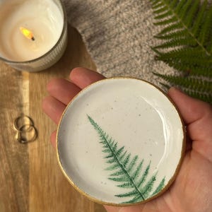 May include: A small, round, white ceramic trinket dish featuring a hand-painted green fern leaf design on the interior surface. The circular dish has a smooth, glossy finish with subtle speckles and a metallic gold painted rim along the edge.