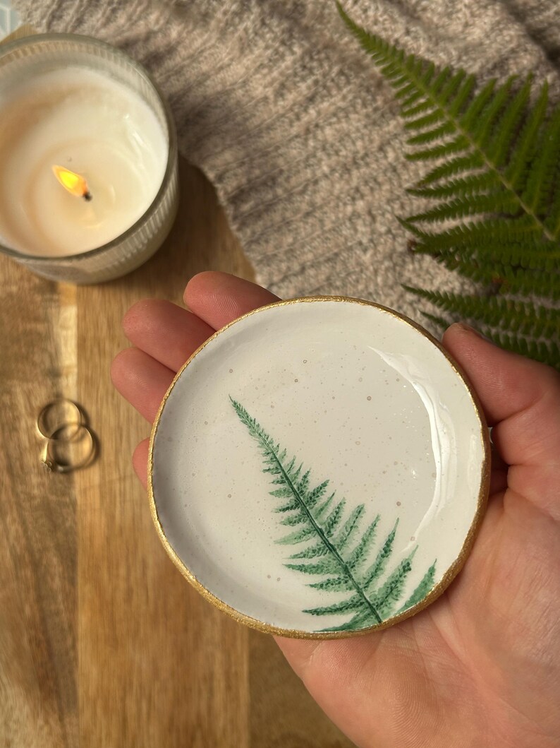 May include: A small, round, white ceramic trinket dish featuring a hand-painted green fern leaf design on the interior surface. The circular dish has a smooth, glossy finish with subtle speckles and a metallic gold painted rim along the edge.