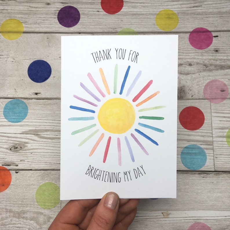 May include: A white card with a watercolour illustration of a yellow sun with rainbow rays. The text "THANK YOU FOR BRIGHTENING MY DAY" is written below the sun.