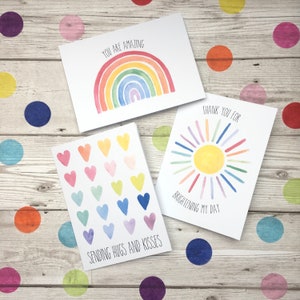 May include: Three greeting cards with watercolour illustrations. The first card features a rainbow with the text "You are amazing". The second card features a sun with the text "Thank you for brightening my day". The third card features a row of hearts in various colours with the text "Sending hugs and kisses".
