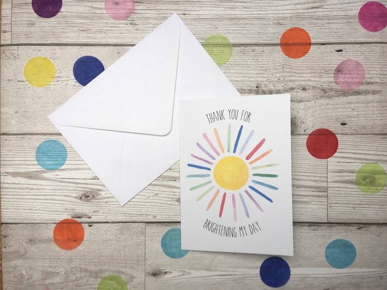 May include: A white card with a colourful sun illustration and the text "Thank you for brightening my day". The card is on a wooden surface with colourful polka dots.
