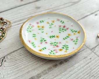 May include: A white ceramic trinket dish with a gold rim and a floral design featuring pink and yellow flowers and green stems.