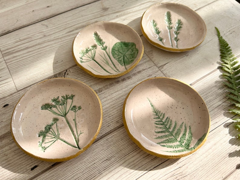 May include: Four round ceramic dishes with a speckled light pink glaze and textured yellow edges. Each dish features a unique green botanical impression, including fern leaves, wildflowers, and a round leaf, creating a nature-inspired set.