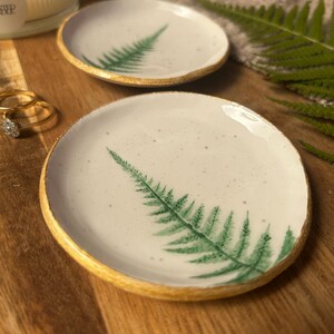 May include: Two round ceramic trinket dishes with a white speckled glaze and a hand-painted green fern leaf design. Each dish features a textured gold-painted rim, providing a metallic finish to the edges of the small, circular clay trays.