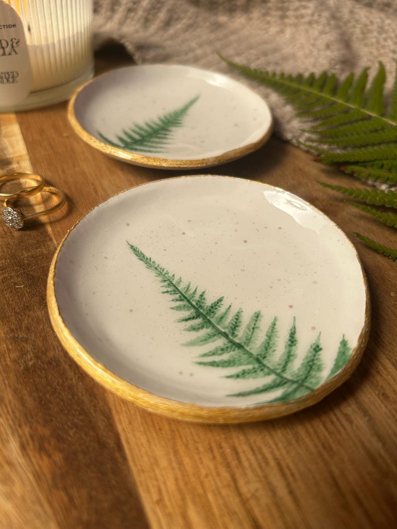 May include: Two round ceramic trinket dishes with a white speckled glaze and a hand-painted green fern leaf design. Each dish features a textured gold-painted rim, providing a metallic finish to the edges of the small, circular clay trays.