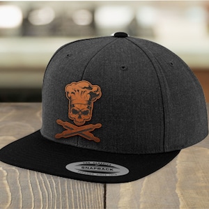 Può includere: Un cappello da baseball nero e marrone con un teschio che indossa un cappello da cuoco e mattarelli incrociati. Il cappello ha una visiera nera e una toppa in pelle marrone con il testo "The Classic Snapback" sul retro.