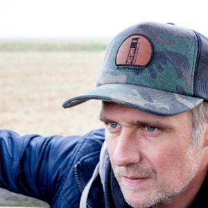May include: A man wearing a camouflage trucker hat with a brown leather patch featuring a hunting blind silhouette and the text "Watchtower" in white. The hat has a black mesh back.