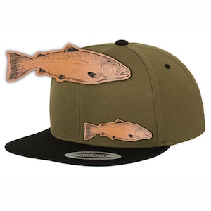 Puede incluir: Una gorra de béisbol verde y negra con dos parches de salmón de cuero marrón. Los parches están cosidos en la parte delantera de la gorra. La gorra tiene una visera negra y una corona verde. El texto "Snapback" está impreso en la parte posterior de la gorra.