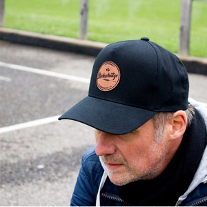 Könnte beinhalten: Eine schwarze Baseballkappe mit einem braunen Lederpatch, auf dem in weißen Buchstaben "Stockhütze" steht. Die Person, die die Kappe trägt, hat einen Bart und schaut nach unten.