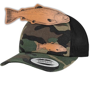 Könnte beinhalten: Eine Camouflage-Trucker-Kappe mit schwarzem Netzrücken. Die Kappe hat einen braunen Leder-Patch mit einer Strichzeichnung eines Lachses. Die Kappe hat einen schwarzen Druckknopfverschluss und den Text "The Classics Snapback" auf der Vorderseite.
