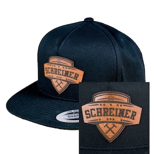 Puede incluir: Gorra de béisbol negra y marrón con un parche de cuero que dice "SCHREINER" con una corona y martillos cruzados.