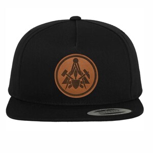 Könnte beinhalten: Eine schwarze und braune Baseballkappe mit einem Lederpatch, der einen Kompass und Werkzeuge zeigt. Der Patch ist rund und hat einen braunen Lederhintergrund.