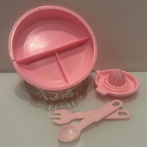Puede incluir: Un juego de utensilios de cocina de plástico rosa claro que incluye un plato redondo dividido, un accesorio para exprimir cítricos, un tenedor pequeño y una cuchara pequeña. El plato está sobre un cuenco de cristal transparente texturizado, junto a los demás.