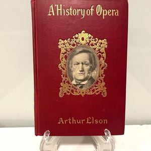 Puede incluir: Libro rojo titulado 'A History of Opera' con letras doradas. La portada presenta un retrato dentro de un marco dorado ornamentado. El nombre del autor, Arthur Elson, también está en oro.