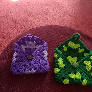 May include: Two handmade crochet envelope pouches. One is purple with a button closure, and the other is green with lime green accents and a button closure. Both are made with a granny square design.