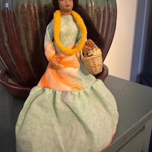 May include: A vintage Hawaiian doll with a floral crown and yellow lei. The doll wears a light green dress with an orange and white bodice and holds a small woven basket. The doll has dark hair and is posed in a seated position.