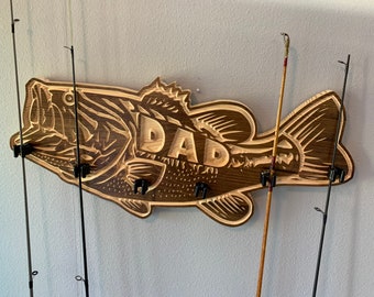 Könnte beinhalten: Wandhalterung aus Holz in Form eines Barsches mit dem Wort "DAD" in die Seite geschnitzt. Die Halterung hat fünf Haken zum Aufhängen von Angelruten. Vier Angelruten hängen an den Haken.