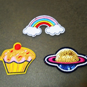 May include: Three embroidered iron-on patches: a rainbow with clouds, a cupcake with pink frosting and red sprinkles, and a gold sequined planet with a pink ring.