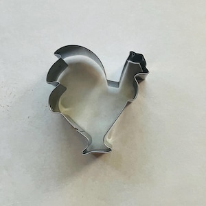 May include: A shiny, silver-colored rooster-shaped cookie cutter. The cutter is made of metal and has a detailed outline of a rooster, including its comb, wattle, and tail feathers. Ideal for baking themed cookies.