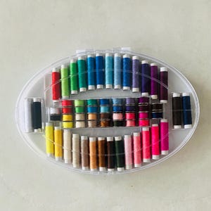 May include: An oval-shaped clear plastic storage case containing a wide variety of sewing thread spools in a spectrum of colors. The spools are arranged in rows and columns within the container, featuring shades of red, yellow, green, blue, purple, pink, brown, black, and white.