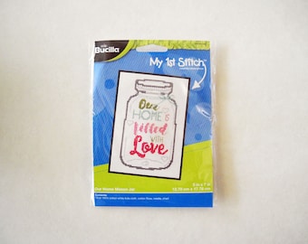 May include: A cross-stitch kit featuring a mason jar with the text "Our Home is filled with Love". The kit is packaged in a blue and white plastic bag with the brand name "Bucilla" and the title "My 1st Stitch". The kit includes 18 count white Aida cloth, cotton floss, needle, and chart. The finished project measures 5 inches by 7 inches.