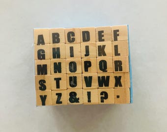 May include: A set of square wooden stamps arranged in a grid, featuring black uppercase letters from A to Z, an ampersand, an exclamation point, a question mark, and a period. Each light-colored wooden block displays a single character clearly.