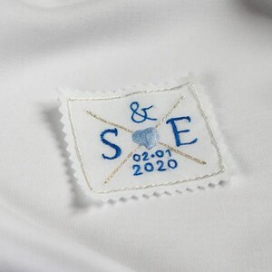 May include: A white fabric patch with blue embroidery. The patch features the letters "S" and "E" with an ampersand symbol between them. A blue heart with a gold outline is centered between the letters. The date "02-01-2020" is embroidered below the heart.